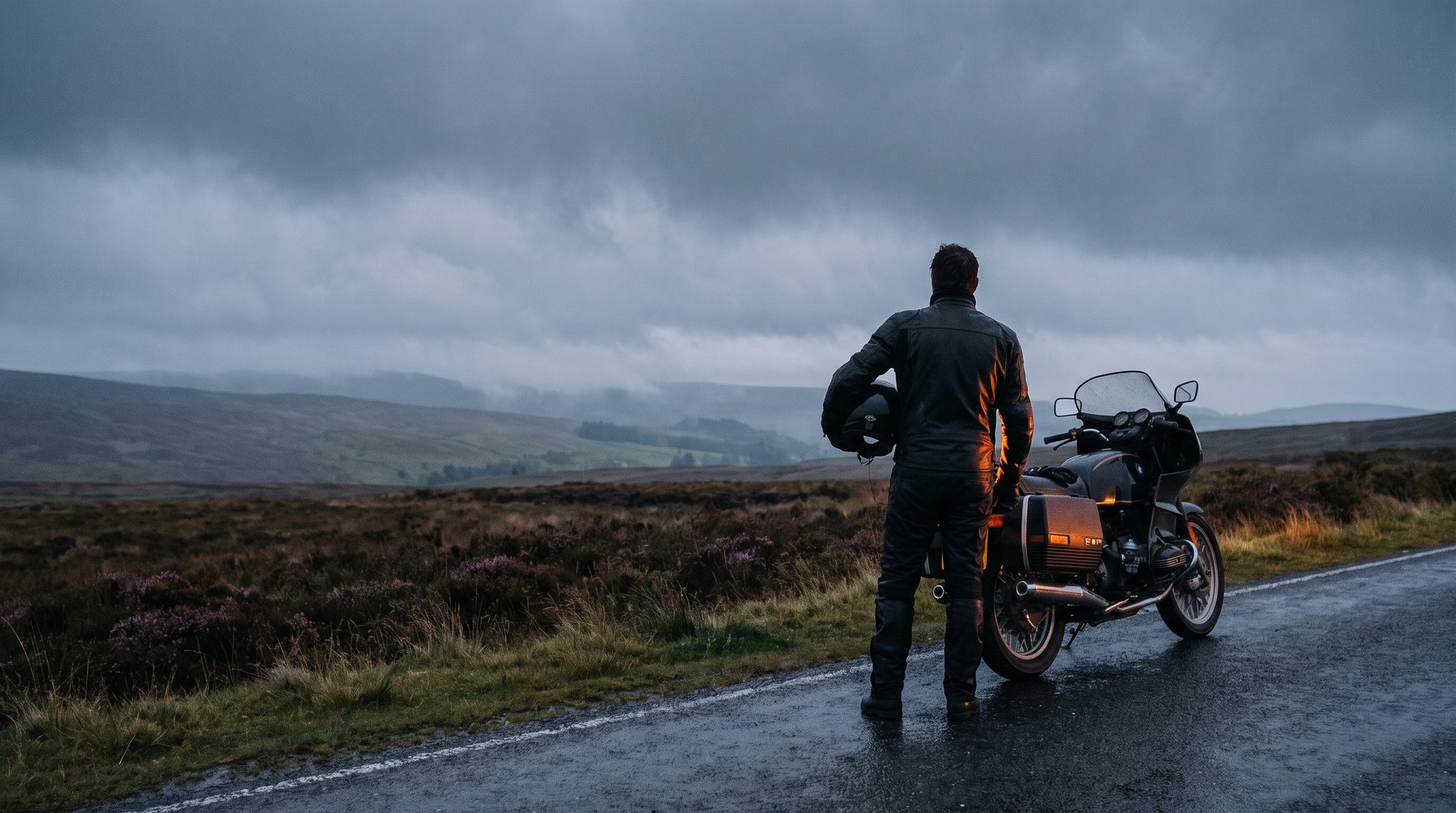 A rider holding a helmet beside a motorcycle on an empty moorland road under heavy cloud.