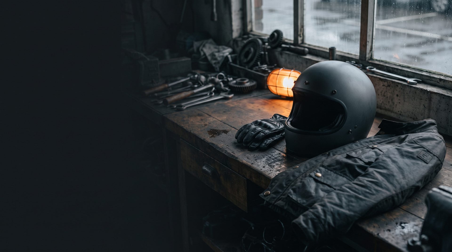 A matt black full face helmet, leather gloves and a waxed jacket on a workbench beside spanners and a lit workshop lamp.